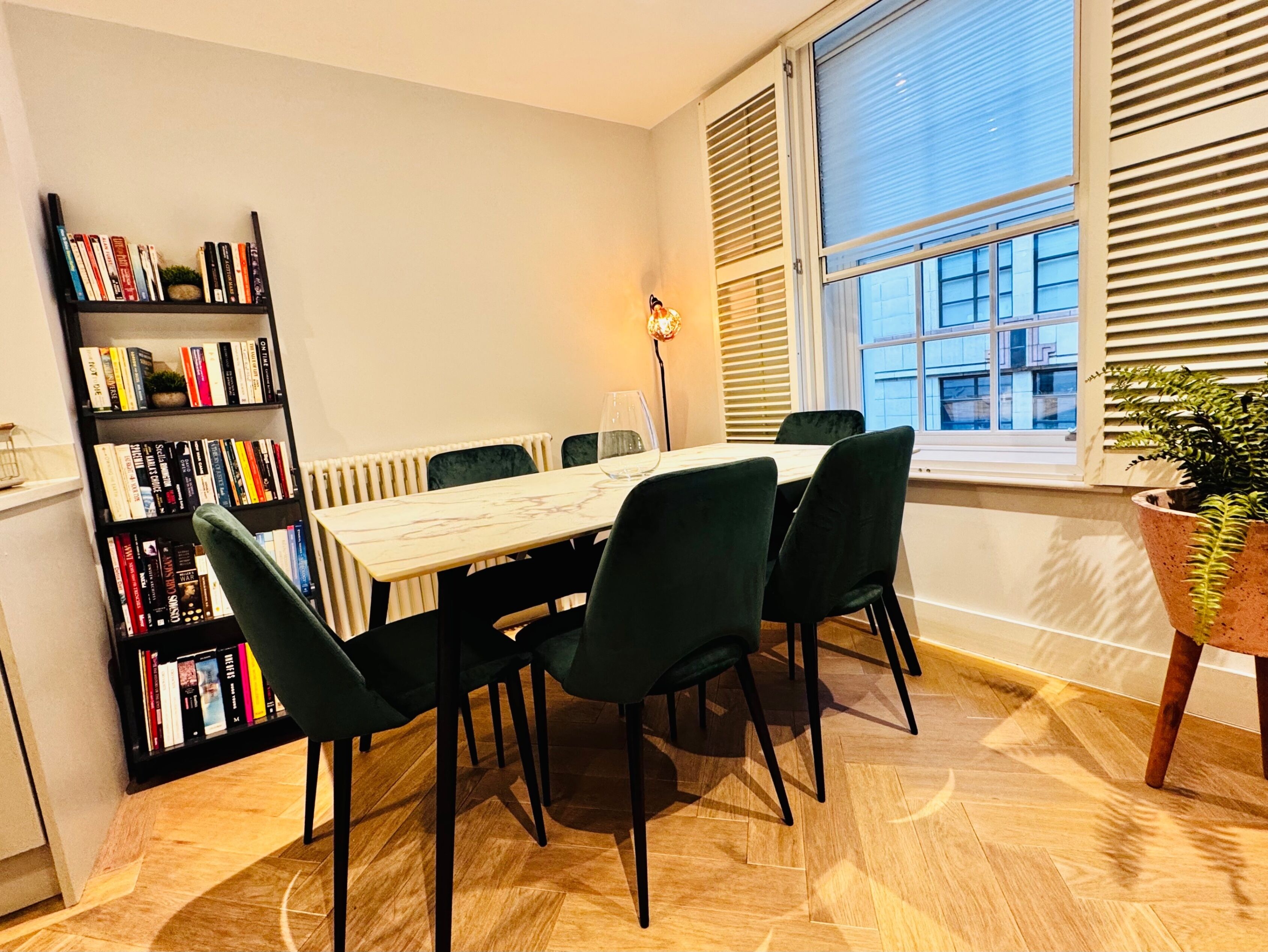 Ein Essbereich mit einem Marmortisch, dunkelgrünen Stühlen, einem Bücherregal und einem Fenster mit Jalousien.