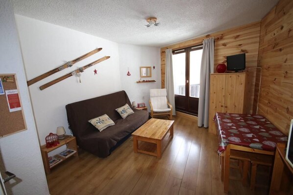 A cozy living room with a brown sofa, wooden coffee table, and a red patterned tablecloth. There is a wooden cabinet with a television, a white chair, and a window with curtains.