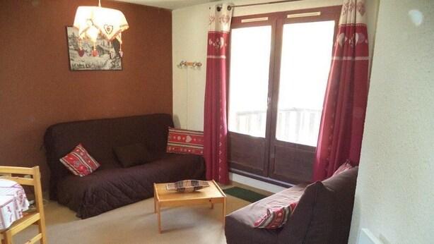 Una sala de estar acogedora con un sofá marrón, una mesa de centro de madera y una silla. Hay una ventana con cortinas rojas y vistas al exterior.