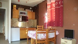 Cocina compacta con armarios de madera, microondas y un refrigerador pequeño. Hay una mesa de comedor con sillas, un televisor sobre un mueble bajo y una cortina roja con motivos de corazones.