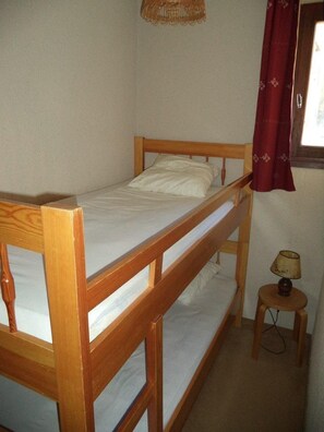A single wooden bunk bed with a mattress and pillows, a small wooden stool with a lamp, and a window with a red curtain.