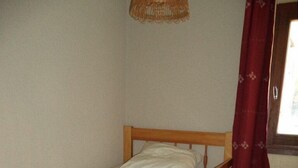 Una litera de madera con colchón y almohadas, un taburete de madera con una lámpara, y una ventana con cortina roja.