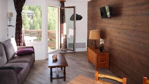 A room with a sofa, a table, a wooden cabinet, a TV mounted on the wall, and a sliding glass door leading to an outdoor area. - 40m from shops, 4th floor, balcony, tv, 30m², Les Orres (Les Orres)