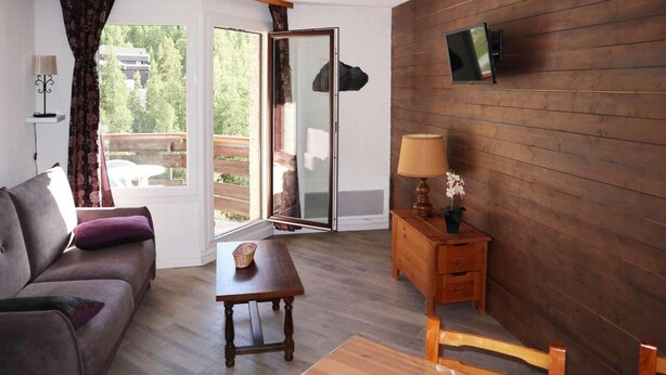 Une pièce comprenant un canapé, une table, un meuble en bois, un téléviseur fixé au mur et une porte-fenêtre coulissante donnant sur un espace extérieur.