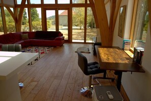 En rummelig stue med trægulv, en rød sofa, et spisebord med stole og et stort vindue med udsigt til det fri.