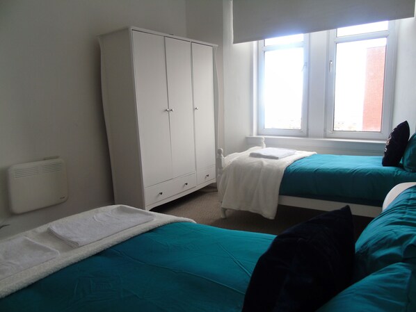 A bedroom with two beds, a wardrobe, a radiator, and a window with a view of a building. - Snug - Grian-aig Apartment (Greenock)