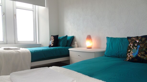 A hotel room with two beds, each with teal bedding and decorative pillows, a white nightstand with a lamp, and a window providing natural light. - Snug - Grian-aig Apartment (Greenock)