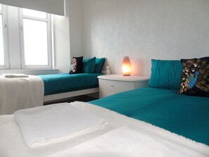 A hotel room with two beds, each with teal bedding and decorative pillows, a white nightstand with a lamp, and a window providing natural light. - Snug - Grian-aig Apartment (Greenock)