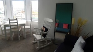 A room with a bay window, a dining table with chairs, a rocking chair, a black sofa, a teal sofa with red cushions, and a mirror. - Snug - Grian-aig Apartment (Greenock)