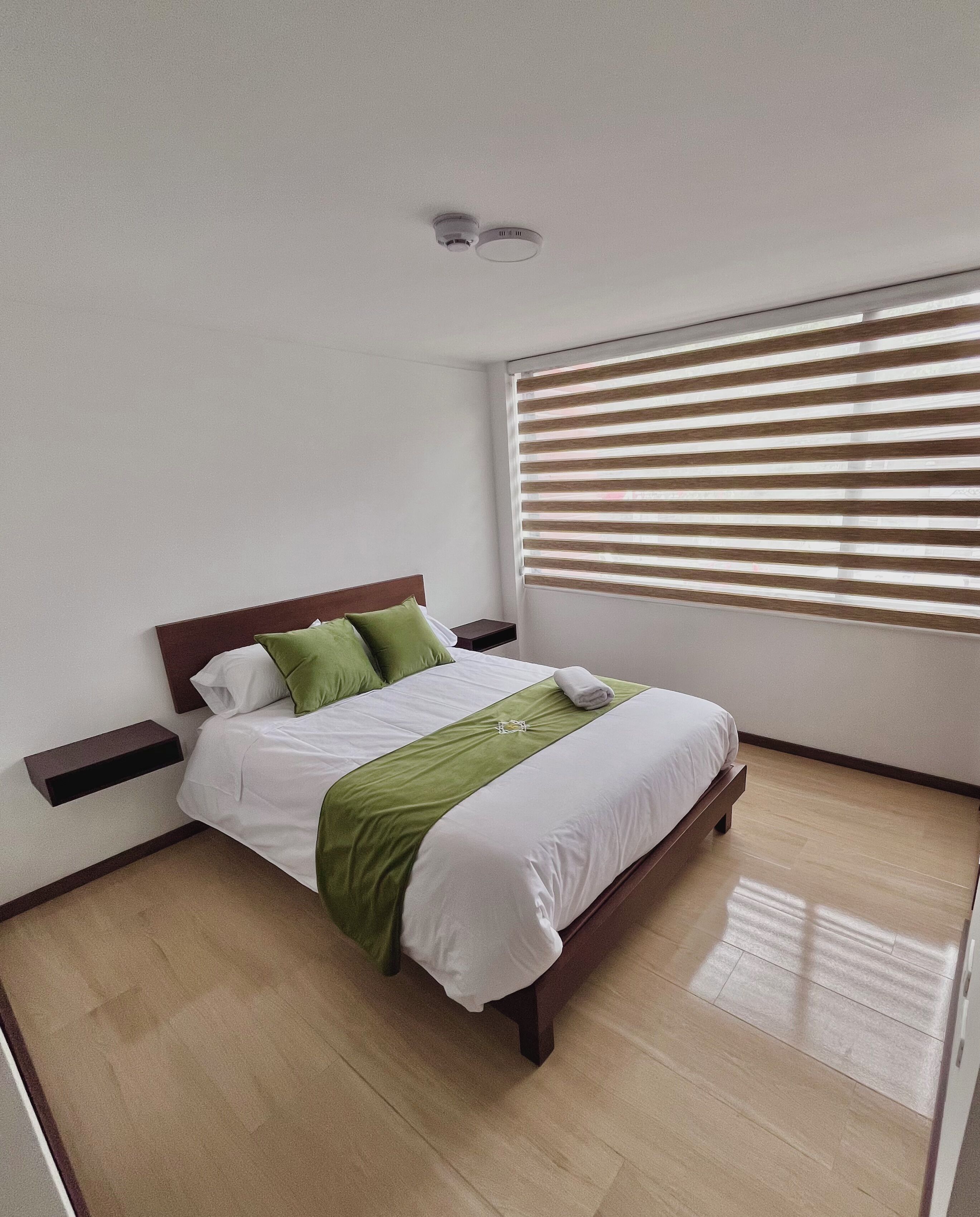 Een netjes opgemaakt bed met een groen-witte dekbedovertrek, een houten hoofdbord en een nachtkastje met een klein plankje.