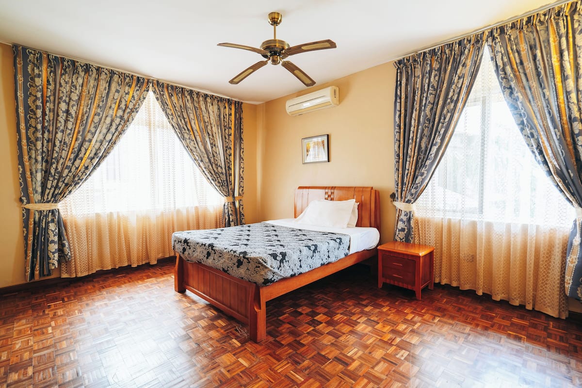 Un dormitorio con cama de madera, mesita de noche, ventilador de techo y ventanas grandes con cortinas.