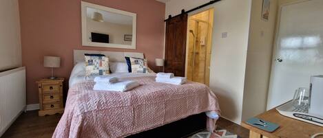 Un letto rifatto con una coperta rosa, un comodino in legno con una lampada, uno specchio grande e una porta scorrevole che conduce a una doccia.