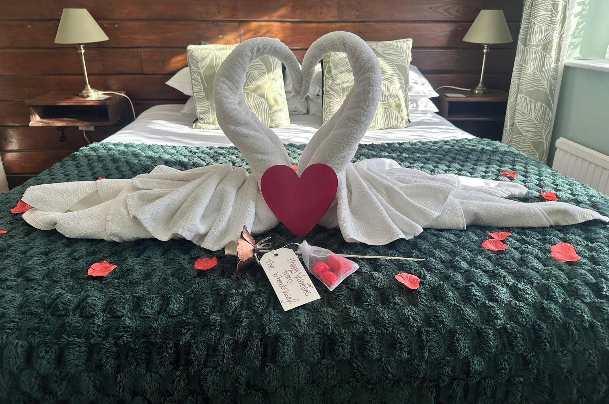 Una cama con una manta verde de punto, una decoración con forma de corazón hecha de toallas y una etiqueta que dice "Feliz Día de San Valentín".