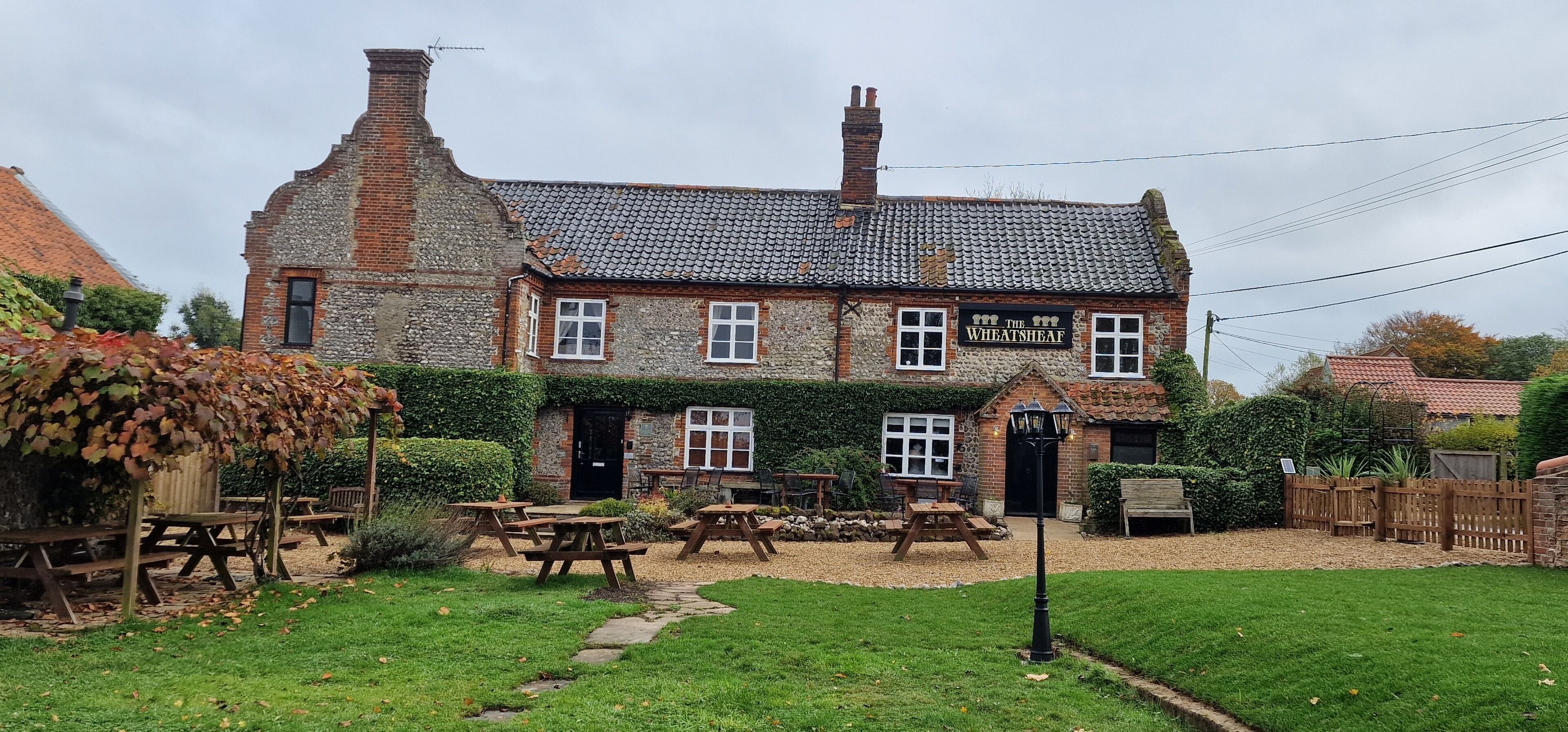 Ein traditionelles Steinhaus mit einem Schild, auf dem "The Wheat Sheaf" steht. Es verfügt über eine Terrasse mit Holztischen und -bänken, umgeben von einem gepflegten Rasen und Garten.
