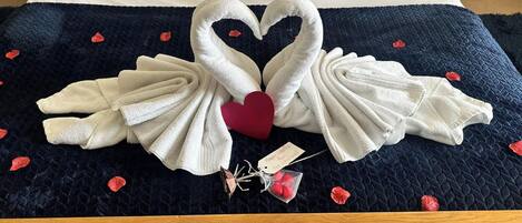 A bed with a dark blue quilt and white towels arranged in the shape of swans with a red heart in the center.