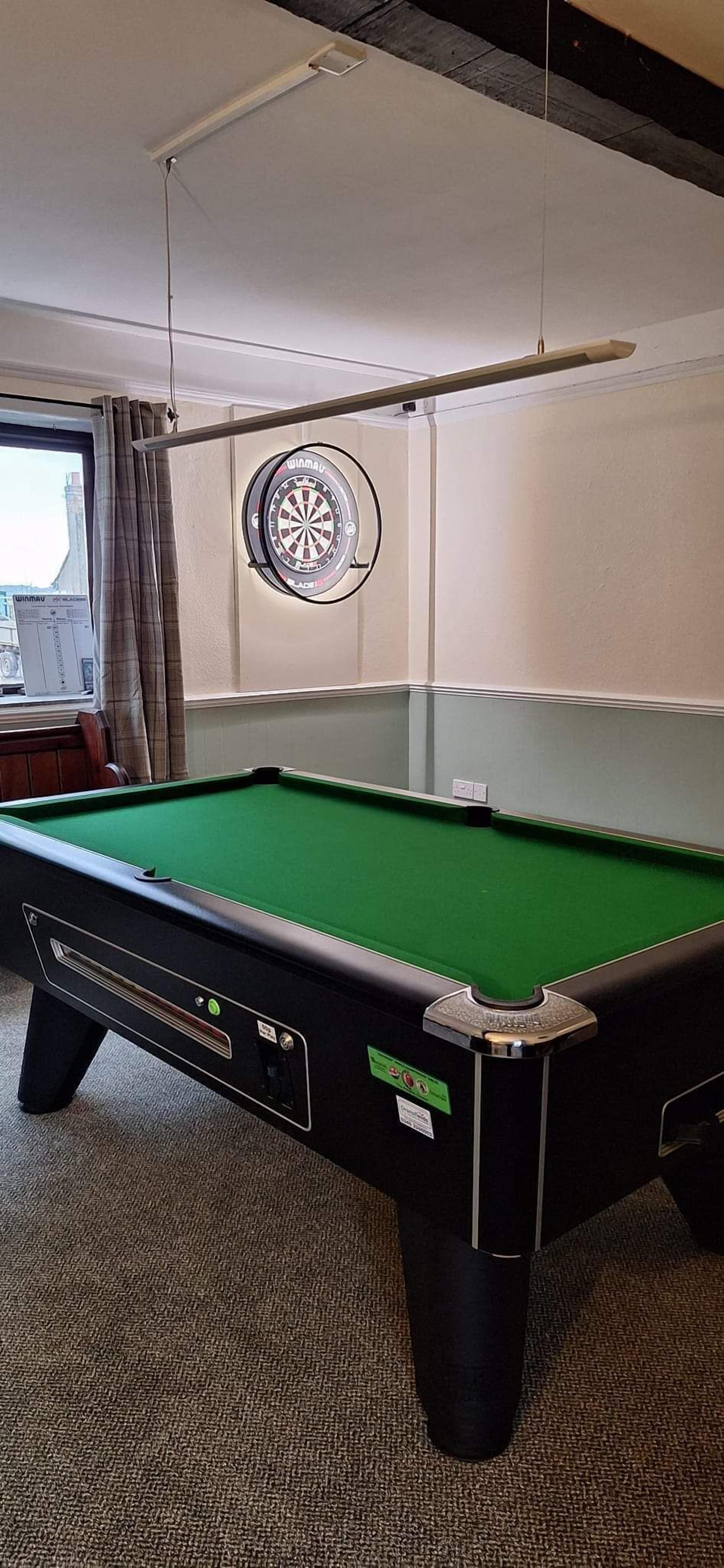 A pool table with a dartboard, carpeted floor, and a window with curtains.