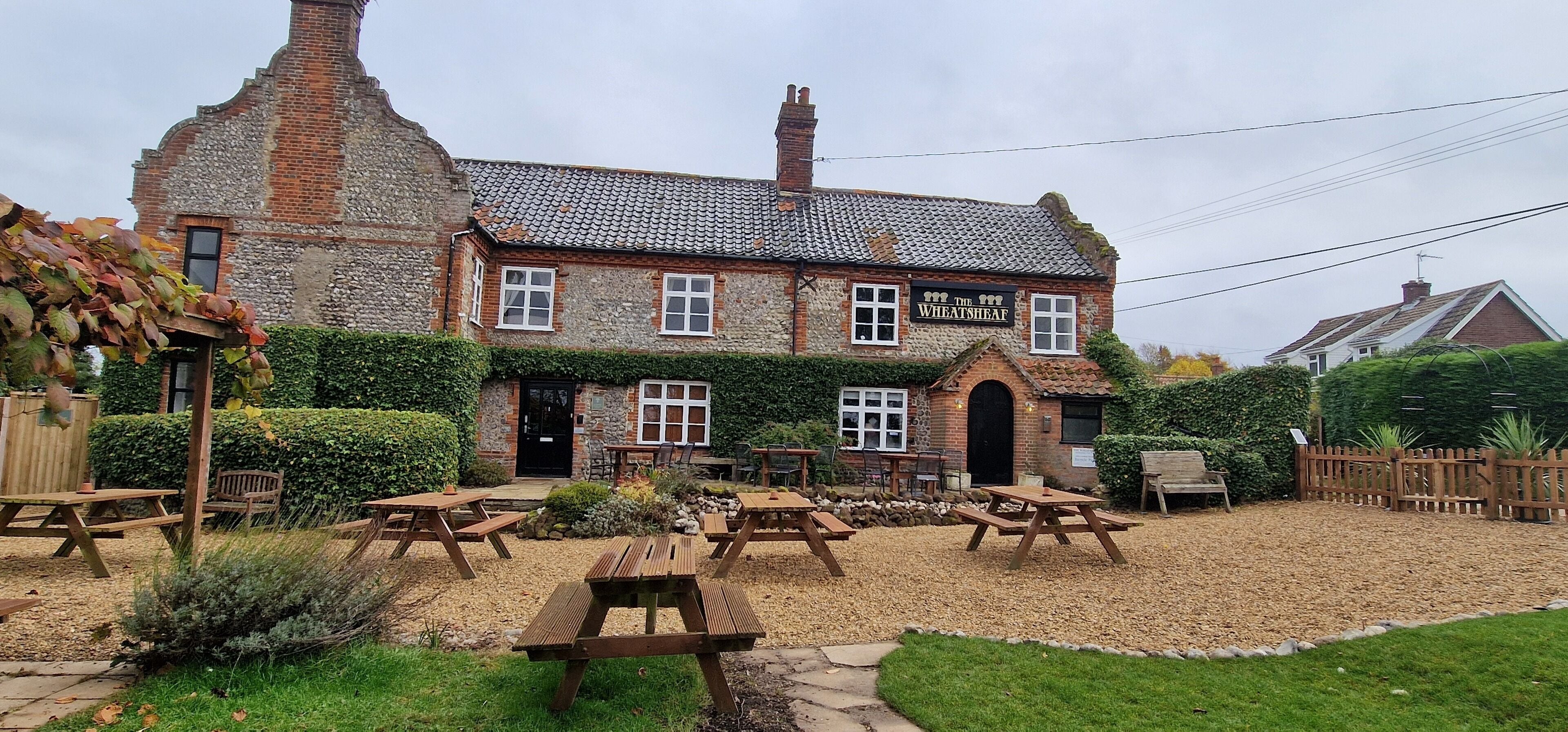 A traditional brick building with a sign reading "The Wheatsheaf." It features a patio with wooden tables and benches, surrounded by a hedge and gravel.