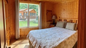 A wooden cabin bedroom with a bed, a nightstand, and a window offering a view of a wooden house and a grassy yard. - Cabin 19 - 1 Bedroom with spacious loft (Isle)