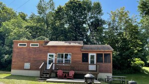 A wooden cabin with a deck, two chairs, a table, a grill, and a chimney. - Cabin 19 - 1 Bedroom with spacious loft (Isle)