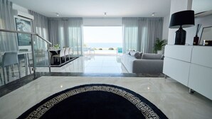 A modern living room with a sofa, a dining area, and a view of the sea.