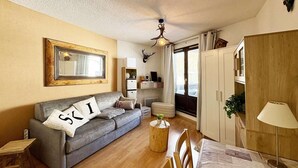 A cozy living room with a grey sofa, a wooden coffee table, and a dining table with chairs. A large mirror hangs on the wall, and a ceiling fan is visible. - 10m from ski slopes, 4th floor, view ski slopes, balcony, tv, 22m², Les Orres (Les Orres)