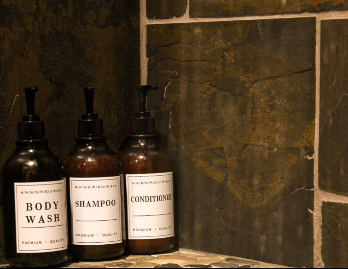 Three bottles of body wash, shampoo, and conditioner on a shelf against a stone wall.