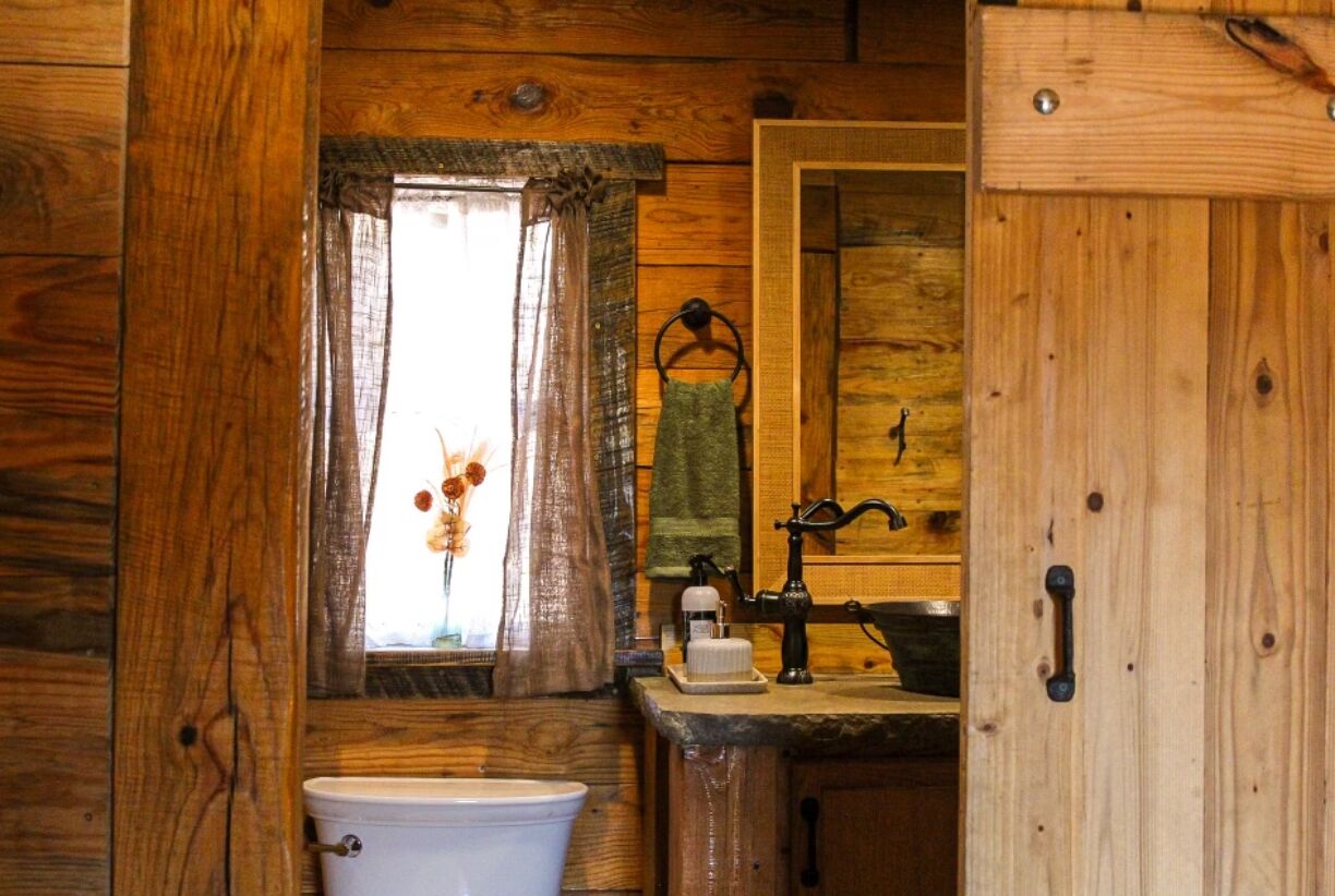 A rustic wooden cabin bathroom with a toilet, sink, and window with a curtain.