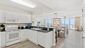 Cucina moderna con mobili bianchi, forno integrato e lavastoviglie. C'è un'area pranzo con tavolo e sedie, e un'ampia finestra con vista sull'oceano.
