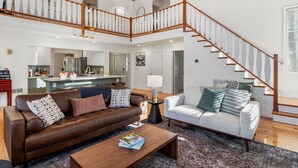 A spacious living area with a brown leather sofa, a white sofa, and a coffee table. A wooden staircase with a railing leads to an upper level.