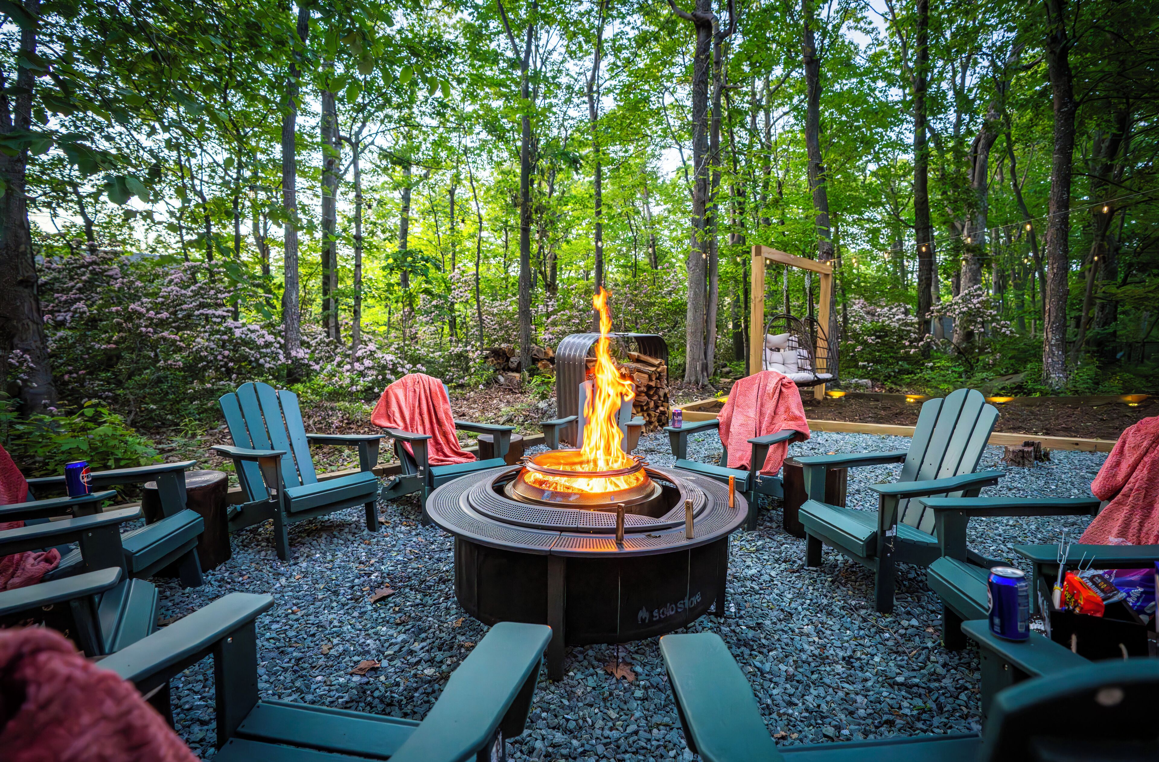A circular fire pit surrounded by green Adirondack chairs with blankets and a can of beer on one of them.