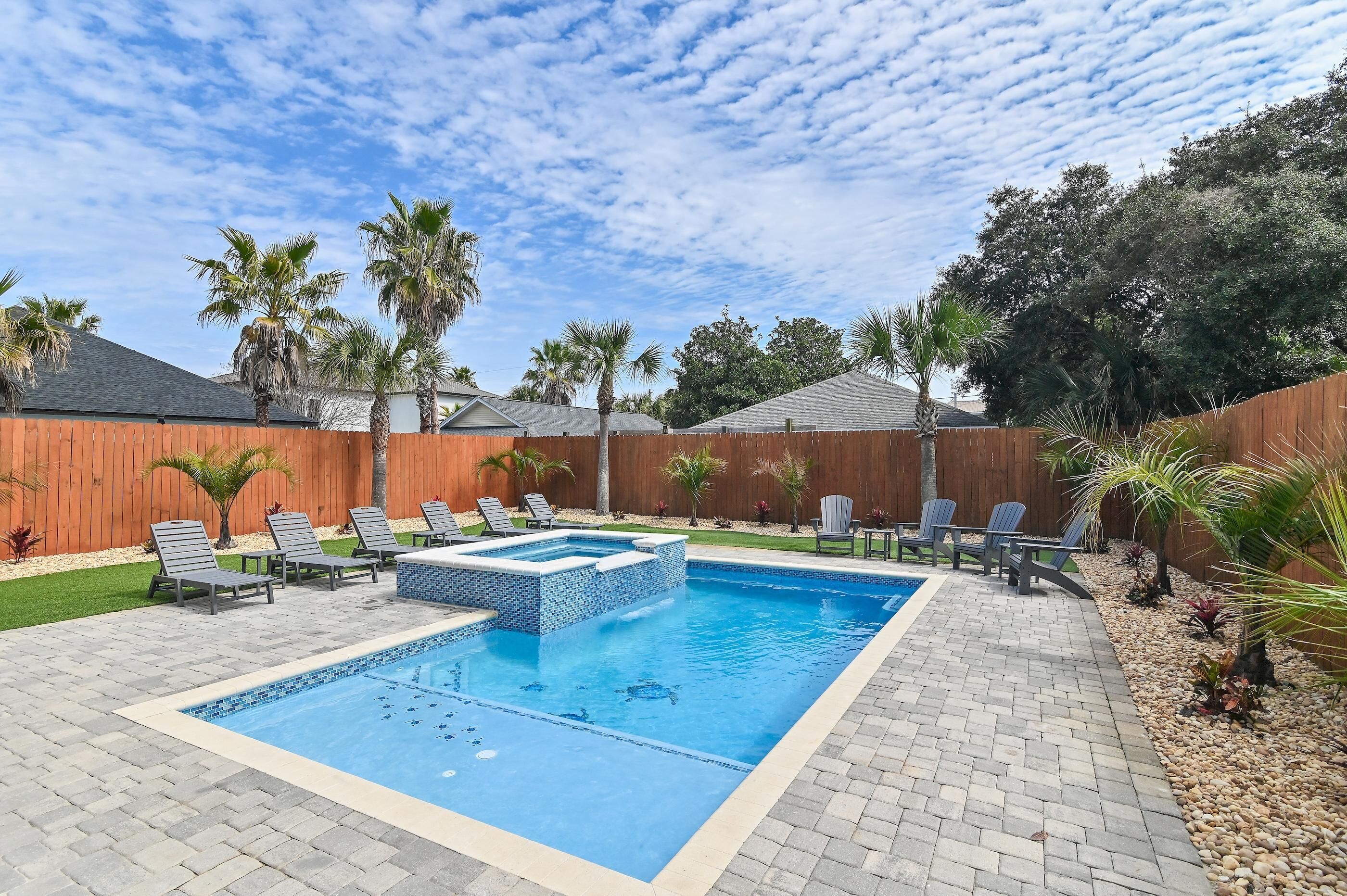 A rectangular swimming pool with a tiled deck, surrounded by lounge chairs and a wooden fence.