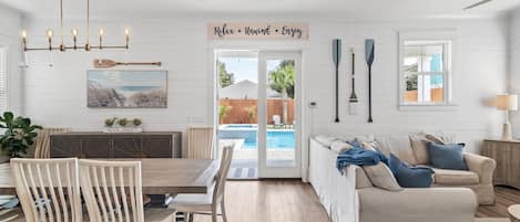 A spacious living room with a wooden dining table, beige sofa, and a view of a swimming pool through glass doors.