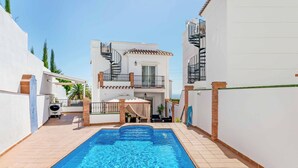A swimming pool with a sun lounger and a table with drinks, surrounded by white walls and a two-story building with a balcony. - Villa Juncos (Nerja)