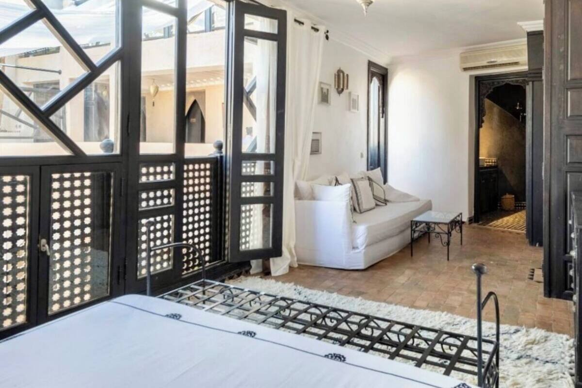 A room with a large window, a white sofa, a small table, and a bed with a white headboard.