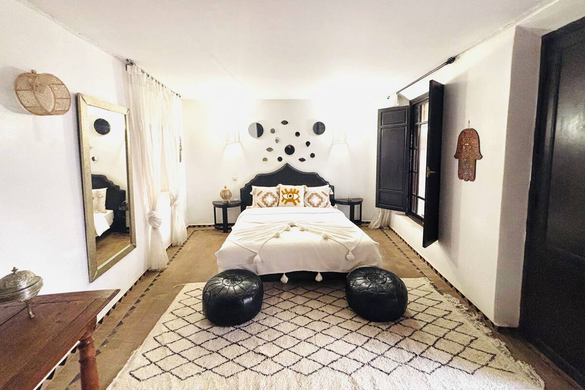 A bedroom with a bed centered in the room, flanked by two nightstands with lamps, a mirror on the left, and a wardrobe on the right.