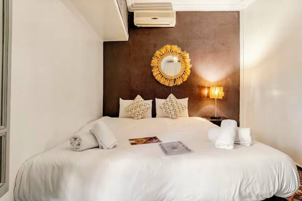 A neatly made bed with white linens, a round mirror, and a wall-mounted lamp.
