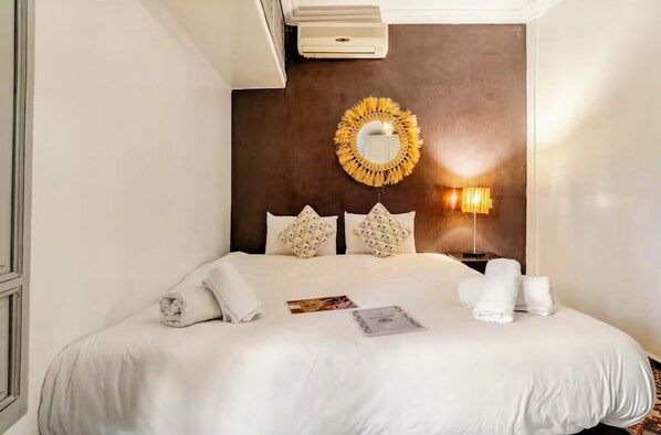 A neatly made bed with white linens, a round mirror, and a wall-mounted lamp.
