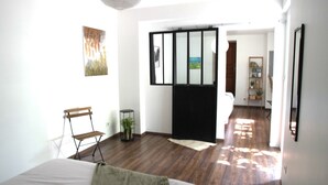 A spacious room with a large bed, a wooden bench, a small round table with a plant, and a sliding glass door leading to another room.