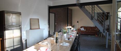 A dining area with a long wooden table set for six, a refrigerator, and a staircase leading to another level.