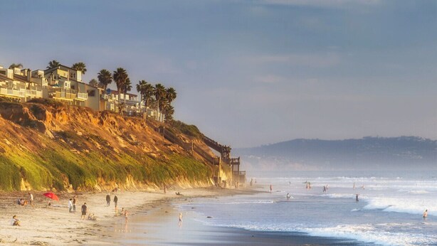 A coastal beach with buildings on a cliff, a lifeguard tower, and people on the sand. - On Omni La Costa Golf Course- Patio, Parking, & AC (Carlsbad)