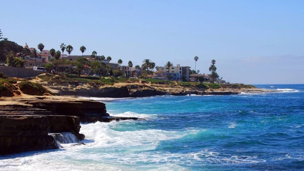 A coastal landscape with rocky cliffs, ocean waves, and a residential area with buildings and palm trees.