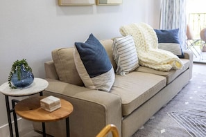 Un canapé beige agrémenté de plusieurs coussins, une petite table ronde en bois sur laquelle repose un objet blanc, et un vase en verre bleu contenant des plantes vertes.