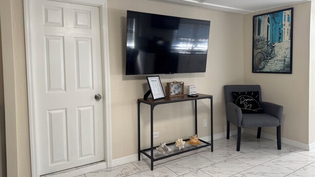 A living room with a white door, a mounted flat-screen TV, a small console table with decorative items, a gray armchair, and a framed picture on the wall. - Cozy 2-bedroom apartment 3 mins away from
Tampa Int’l airport  (Tampa)