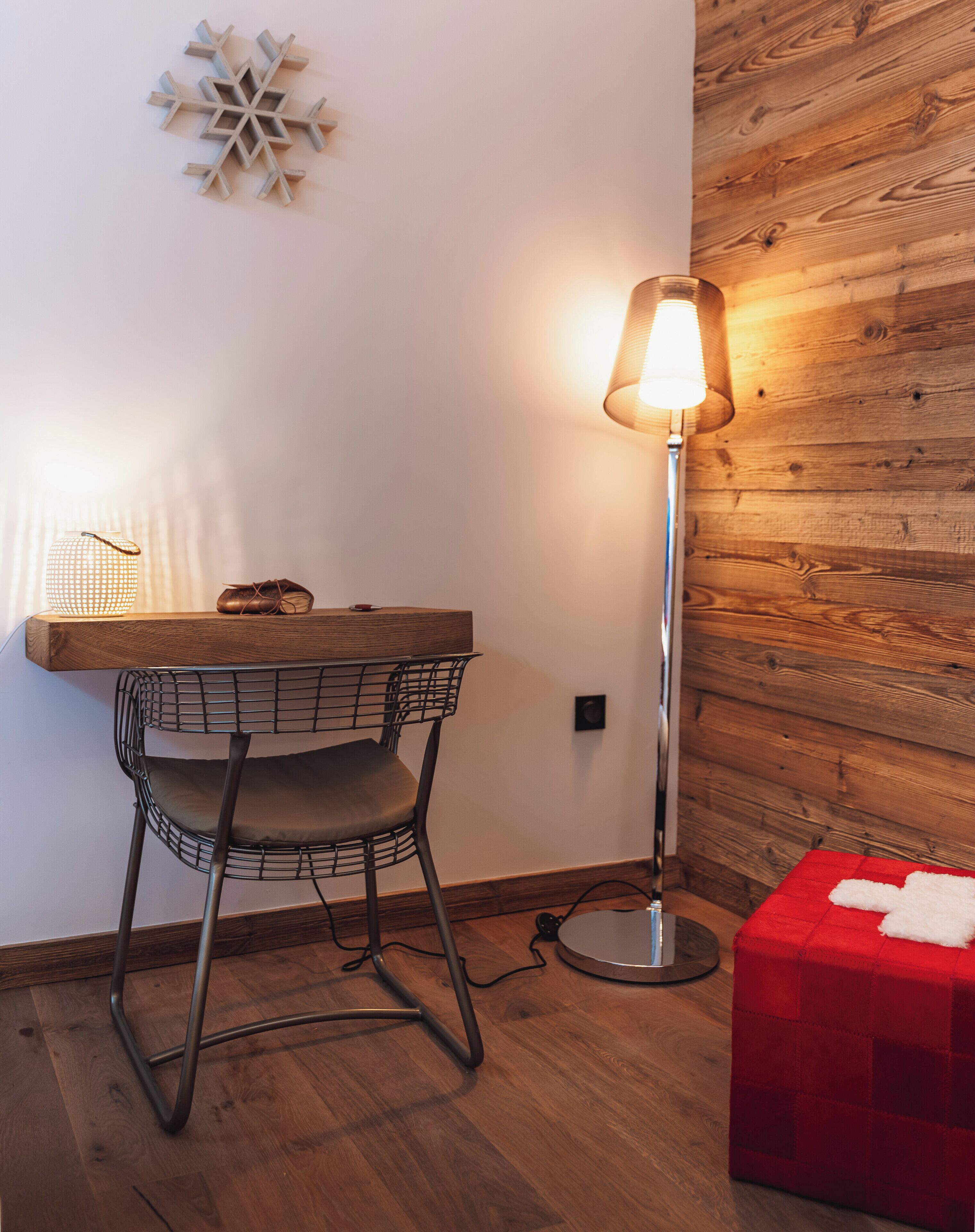 Un suelo de madera, una silla de metal, una mesa, una lámpara de pie y un reposapiés rojo.