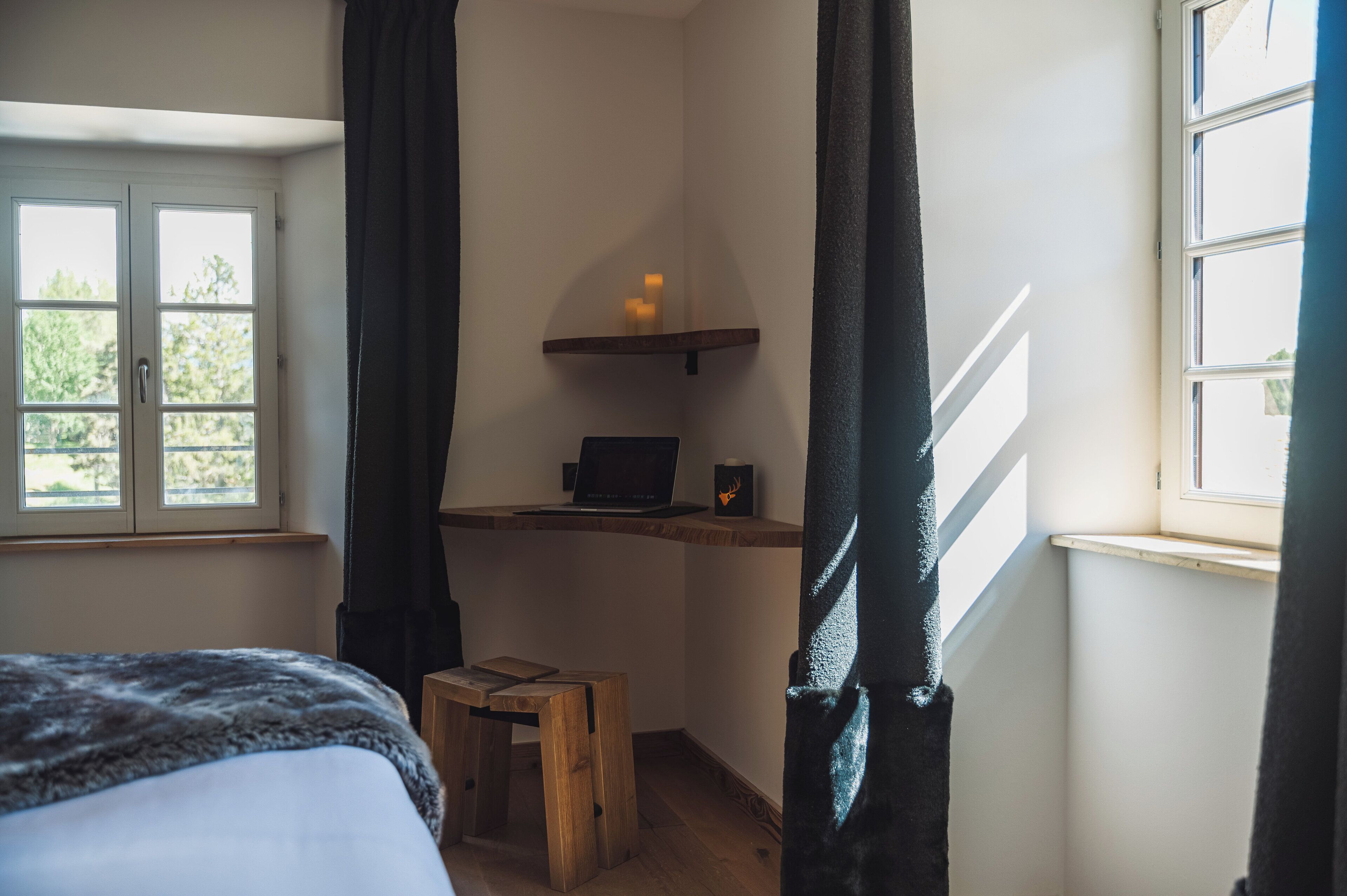 A bedroom with a bed, a wooden stool, a desk with a laptop and candles, and a window with curtains.