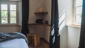 A bedroom with a bed, a wooden stool, a desk with a laptop and candles, and a window with curtains.