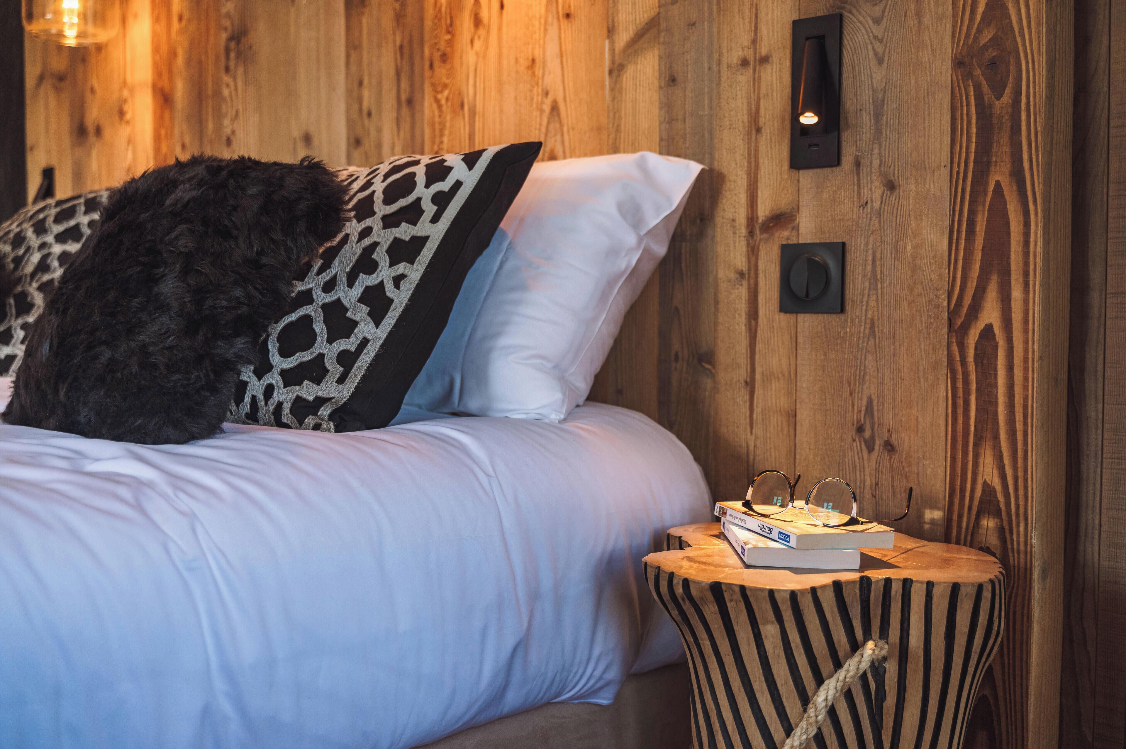 A bed with patterned pillows, a bedside table with books and glasses, and wooden paneling.