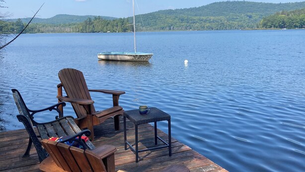 A wooden dock with chairs and a table, overlooking a calm lake with a sailboat and a distant hill.
