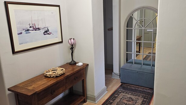 A hallway with a wooden console table, a framed painting, a lamp, and a blue upholstered bench. - Snug - Macushla Apartment (Helensburgh)