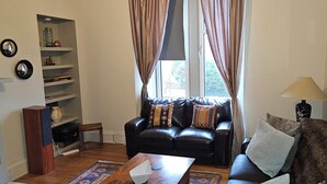 A living room with a black leather sofa, a wooden coffee table, a bookshelf, and a window with curtains. - Snug - Macushla Apartment (Helensburgh)